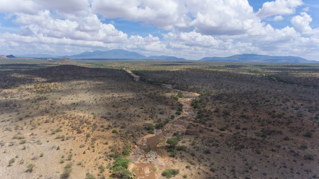 Aerial view of a seasonal river cutting through semi-arid bushland with mountains in the distance, Isiolo County