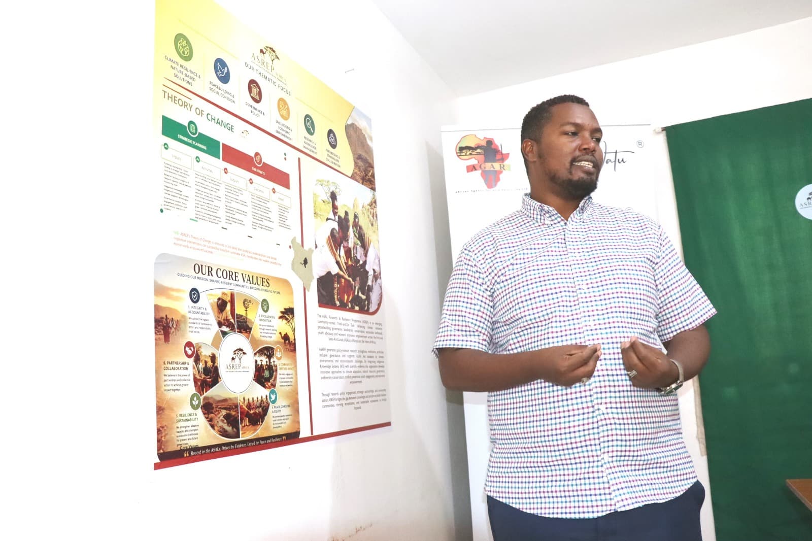 Dida Fayo speaking beside the ASREP thematic-focus and theory-of-change poster and the AGAR banner