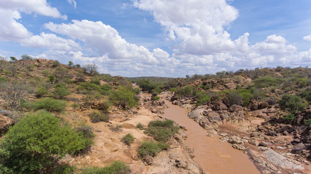 The muddy Ewaso Ny'iro River passing through a rocky gorge with acacia scrub under a big cloud-filled sky